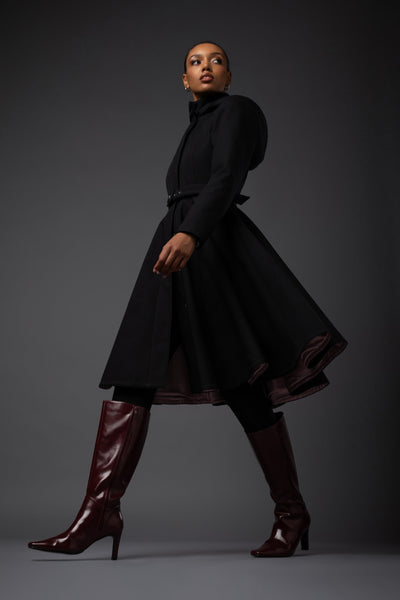 A model in a Rainsisters waterproof black long-sleeve winter coat with a high collar and flared skirt, paired with dark brown knee-high heeled boots, posed against a dark gray studio background.