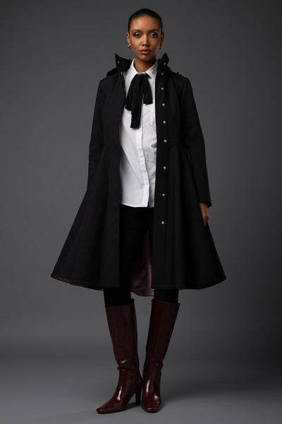 A model stands against a dark gray studio background wearing a long black warm winter coat over a white collared shirt with a black bow, black pants, and dark burgundy knee-high boots.