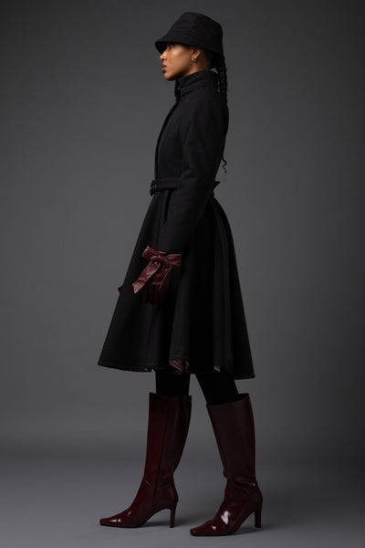 A model in profile wearing a Rainsisters black mid-length belted winter coat, a black hat, black leggings, and dark burgundy knee-high heeled boots against a dark gray studio background.
