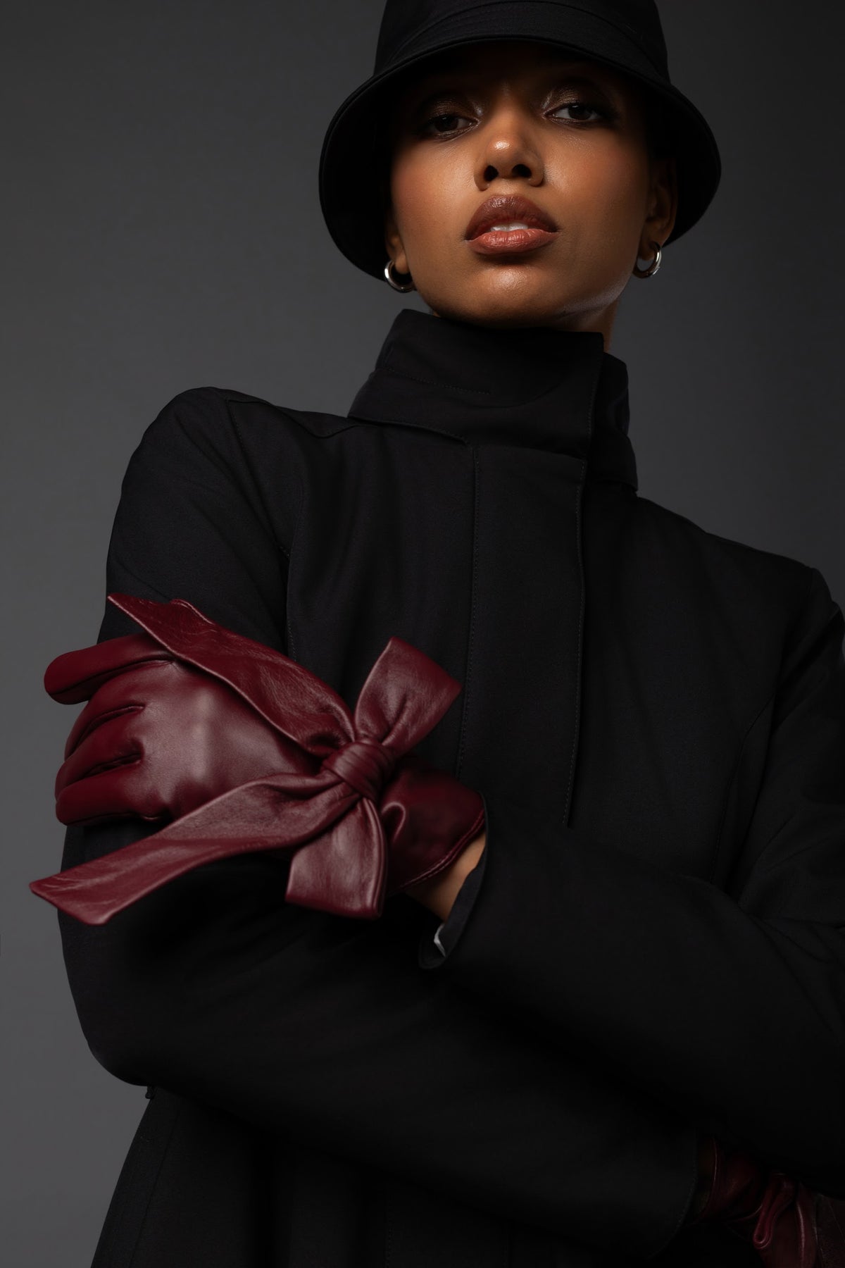 A model wearing a black high-neck coat, black hat, and dark burgundy leather gloves with a large bow detail on the forearm, posed against a dark gray studio background.