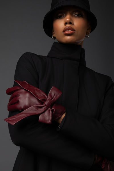 A model wearing a black high-neck coat, black hat, and dark burgundy leather gloves with a large bow detail on the forearm, posed against a dark gray studio background.
