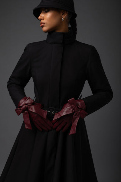 A model wearing a black high-neck long-sleeve waterproof winter coat with a matching black bucket hat, paired with dark burgundy leather gloves, posed against a dark gray studio background.