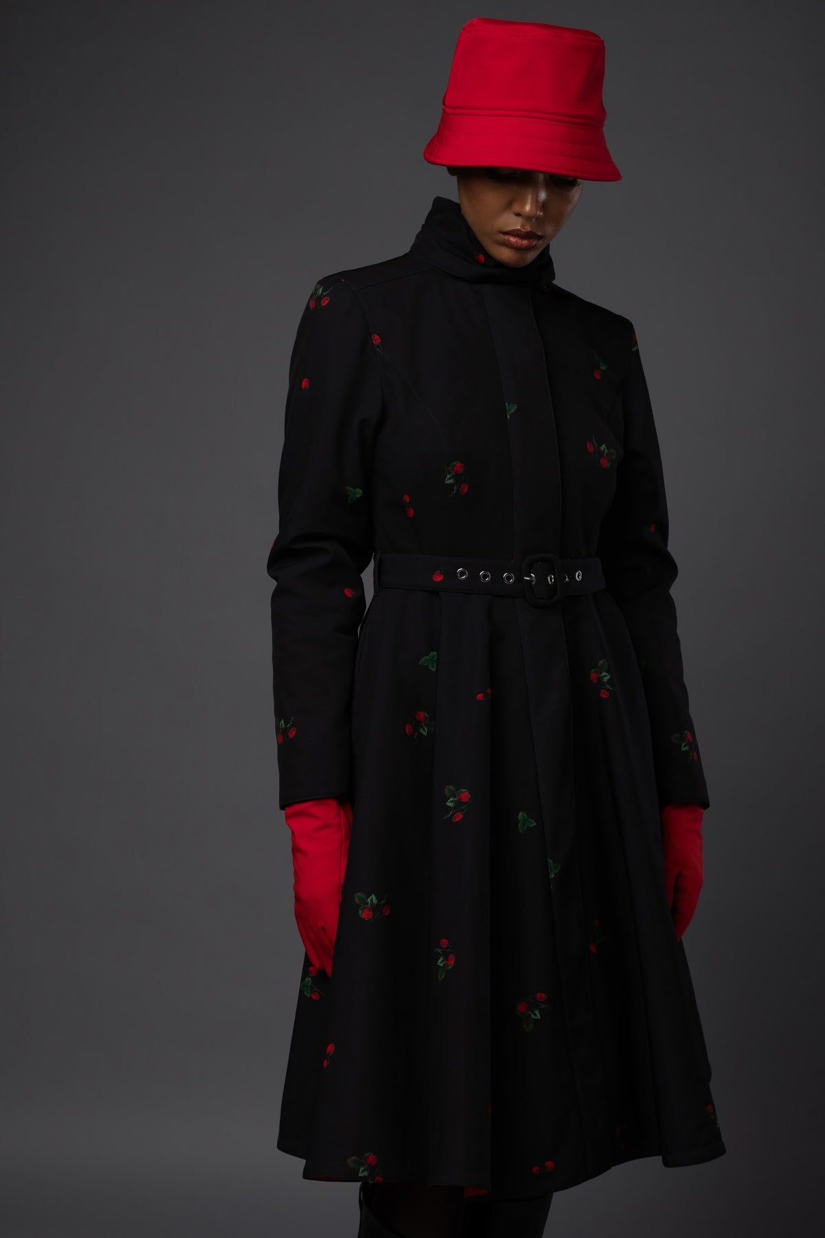 A model wearing a long black winter coat with small red and green berry motifs, styled with a matching red bucket hat, red gloves, and a black belt with silver rings, set against a dark gray studio background.