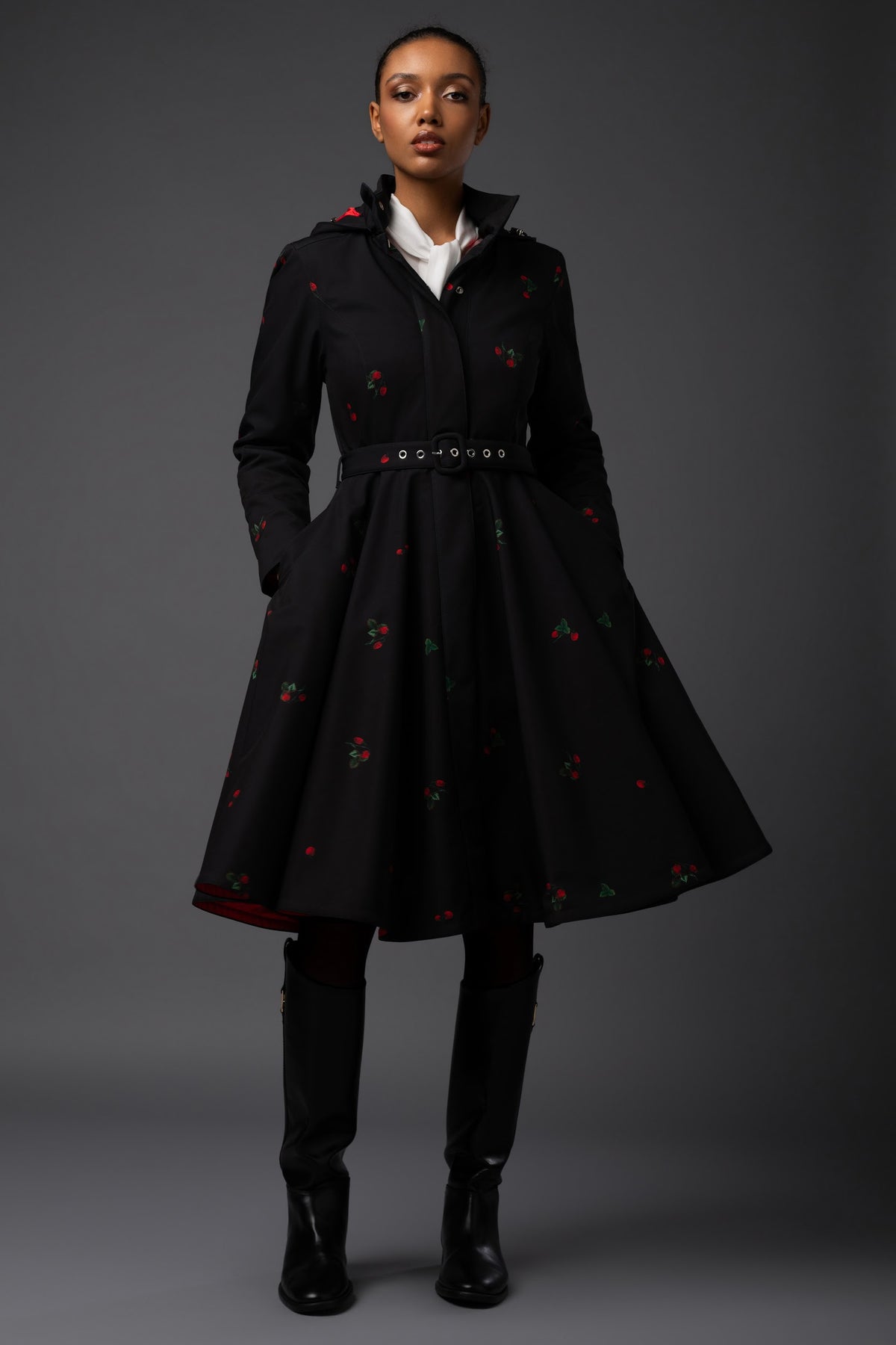 A model stands against a dark gray studio background wearing a black A-line coat with delicate red berry print and green floral motifs, styled with a white collared shirt, a black belt, and black knee-high boots.