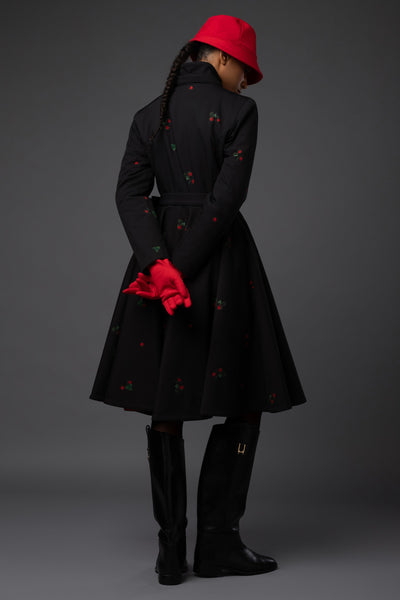 A model shown from the back wearing a black winter coat with small red and green print, a bright red bucket hat, red gloves, and black boots against a dark gray studio background.