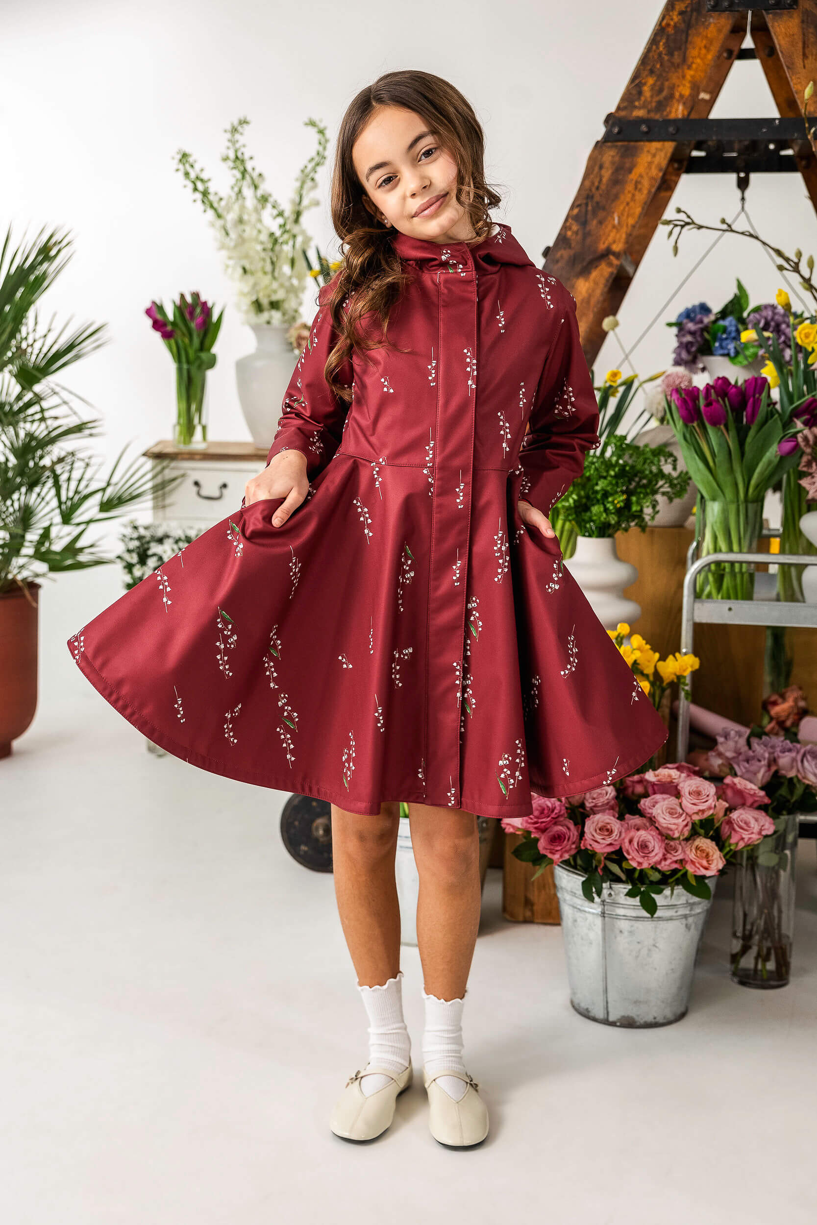 Young girl in a red dress a like coat with floral pattern standing in a room with flowers.