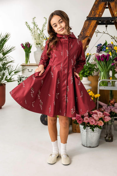 Young girl in a red dress a like coat with floral pattern standing in a room with flowers.