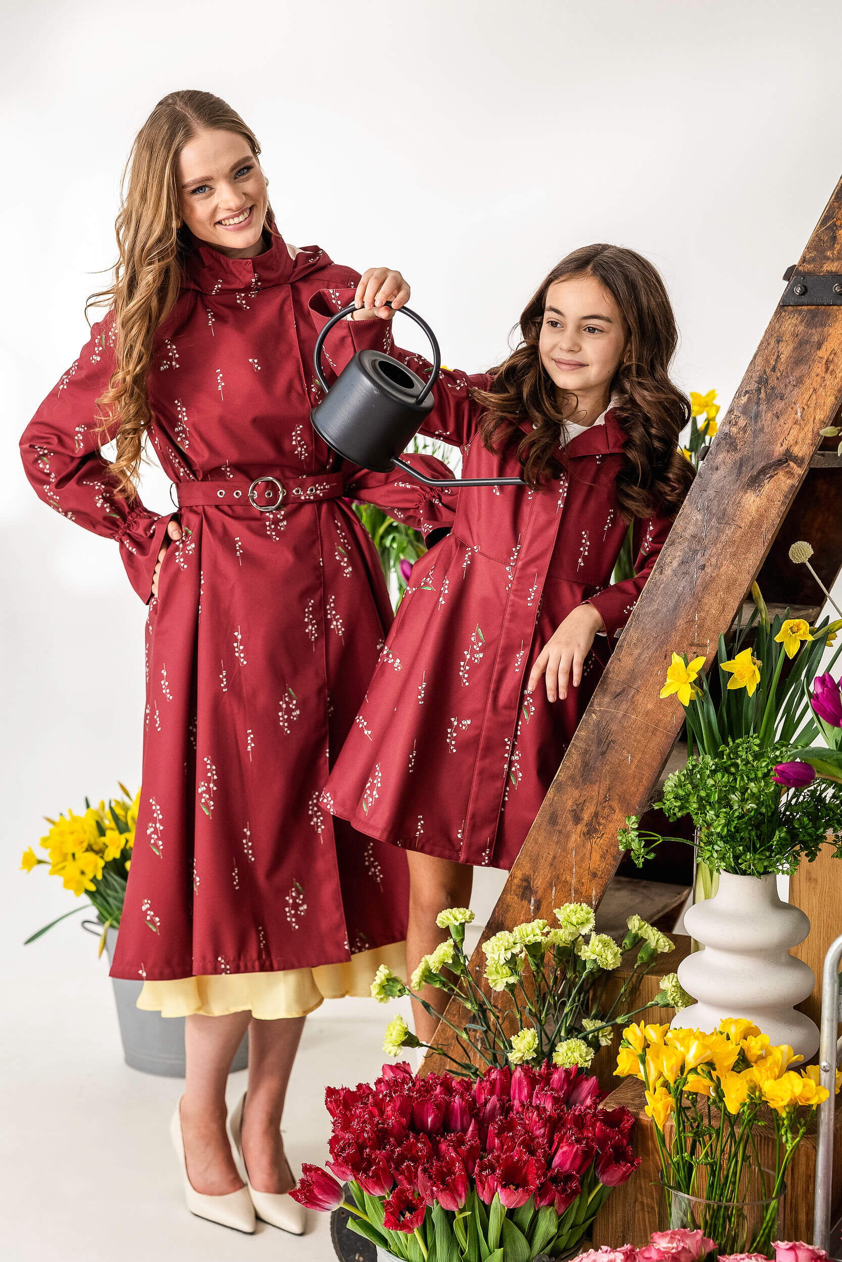 Two women in matching red floral raincoats standing next to a wooden ladder with flowers around them.